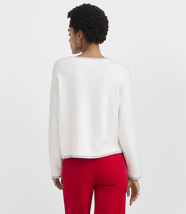 A woman with short curly hair wearing a white sweater and red pants stands against a plain light gray background.