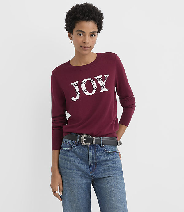 A woman wearing a maroon "JOY" sweater and blue jeans stands against a plain white background.