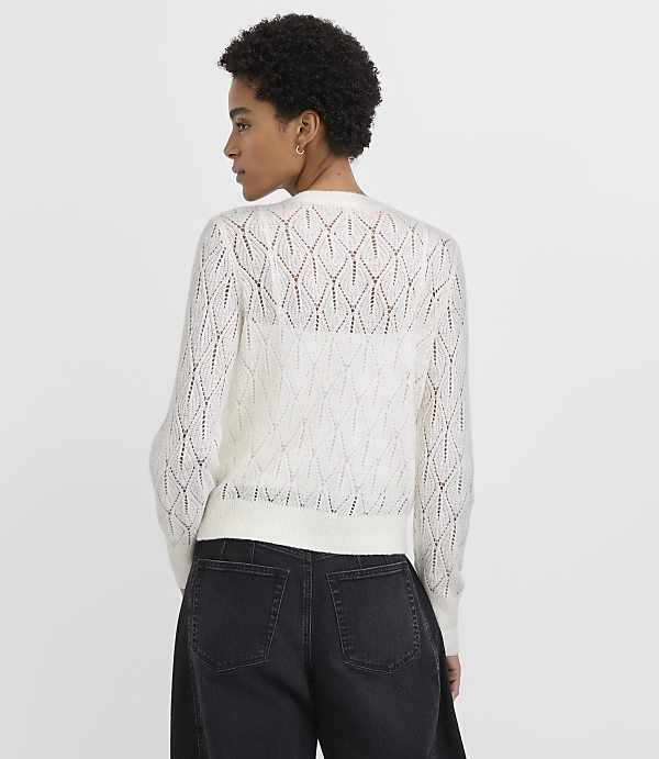 A woman wearing a white open-knit sweater and black jeans stands against a plain light gray background.