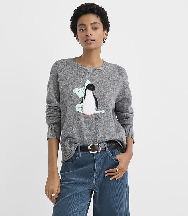 A woman wearing a grey penguin sweater and blue corduroy pants stands against a plain white background.