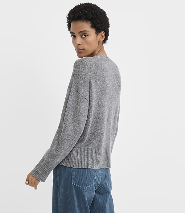 A woman wearing a grey sweater and blue corduroy pants stands against a plain white background.