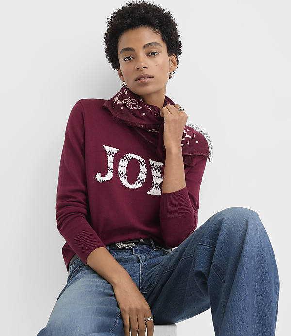 A woman wearing a burgundy "JOY" sweater and blue jeans poses against a plain white background.