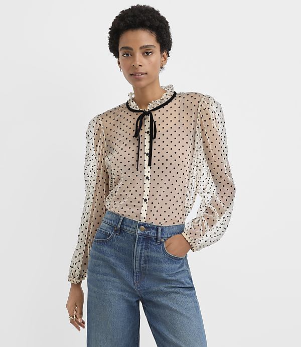 A woman wearing a sheer polka-dot blouse and blue jeans stands against a plain white background.