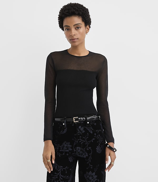 A woman wearing a sheer-sleeved black top and floral patterned black pants stands against a plain white background.