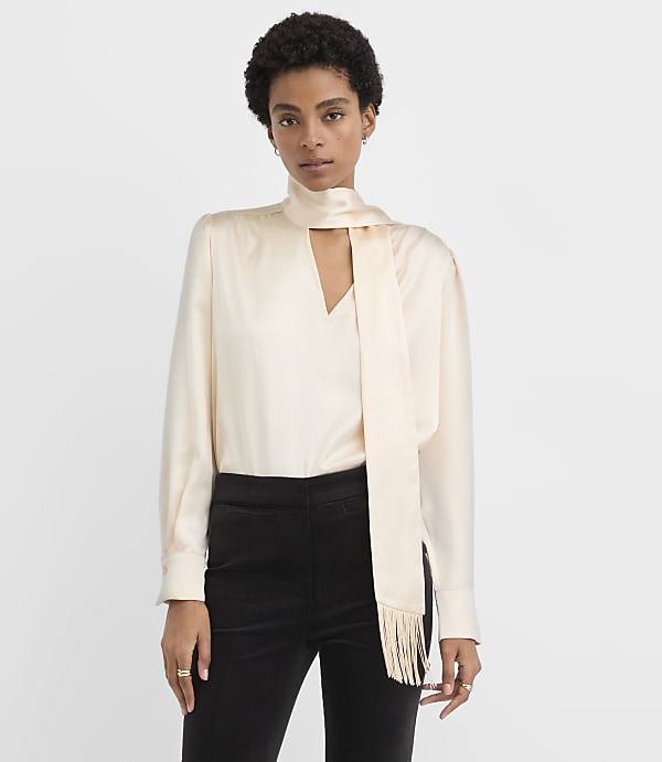 A woman wearing a cream-colored satin blouse with a neck scarf and black trousers stands against a plain white background.