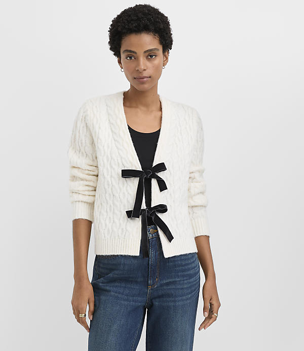 A woman wearing a white cable-knit cardigan with a black bow and blue jeans stands against a plain white background.