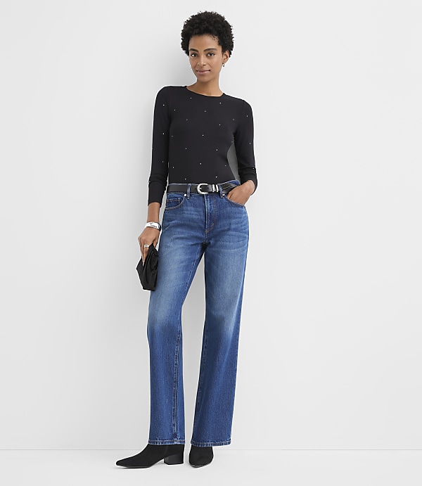 A woman wearing a black studded top, blue jeans, and black boots stands against a plain white background.