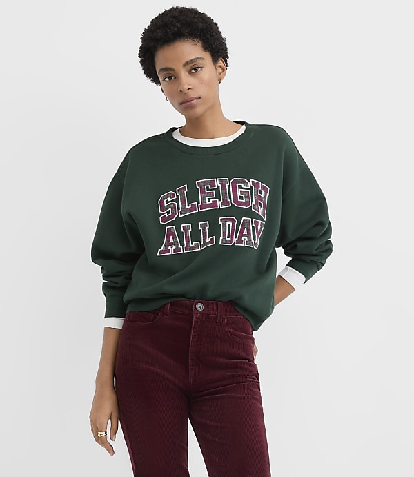A woman wearing a green "SLEIGH ALL DAY" sweatshirt and burgundy corduroy pants stands against a plain white background.