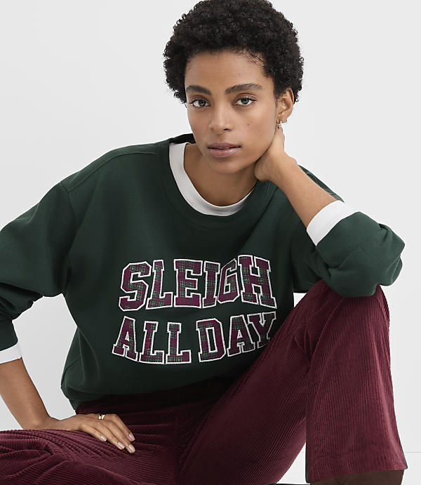 A woman wearing a dark green "SLEIGH ALL DAY" sweatshirt and maroon corduroy pants poses against a plain white background.