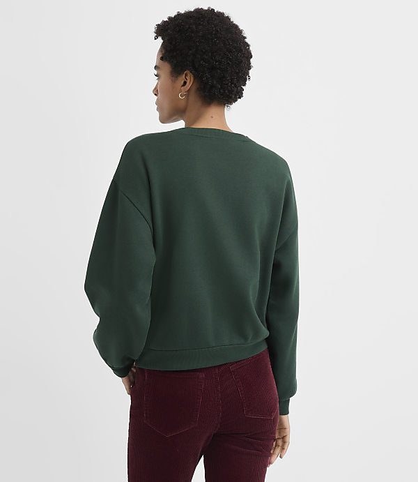 A woman with curly hair wearing a dark green sweatshirt and maroon corduroy pants stands against a plain white background.