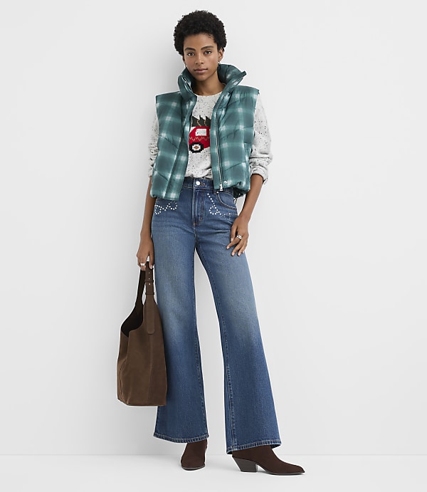 A woman wearing a green puffer vest, a graphic sweater, and flared jeans stands against a plain white background while holding a brown suede tote bag.