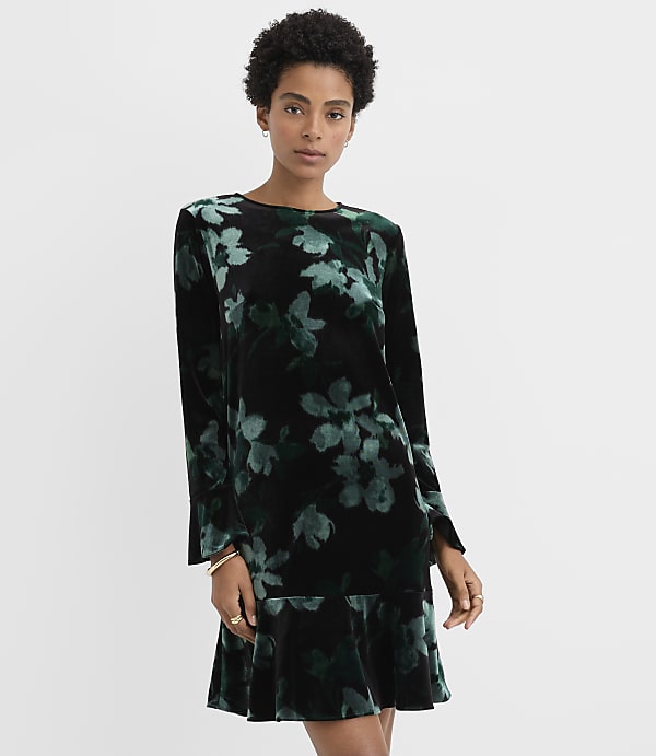 A woman wearing a black and green floral velvet dress stands against a plain white background.