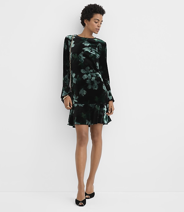 A woman wearing a black and green floral velvet dress and black heels stands against a plain white background.