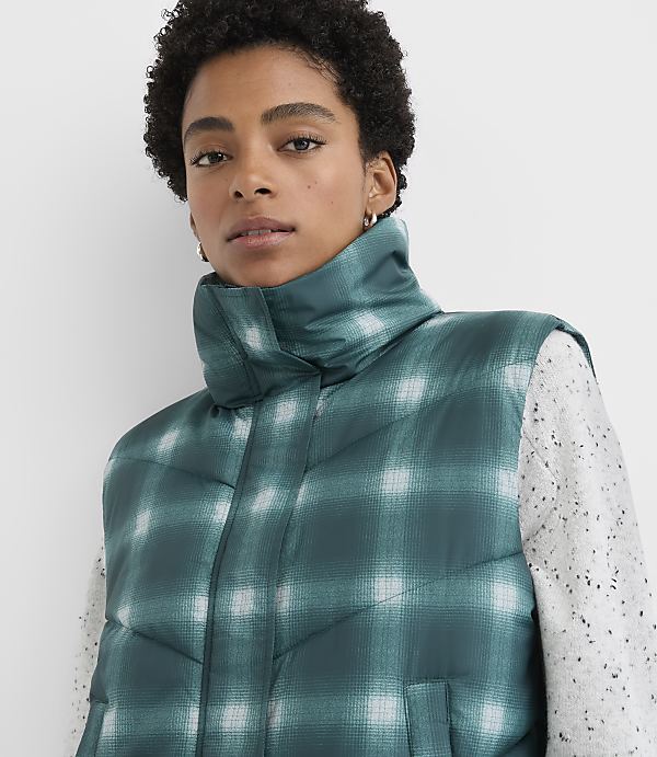 A woman wearing a green plaid puffer vest over a white speckled sweater stands against a plain white background.