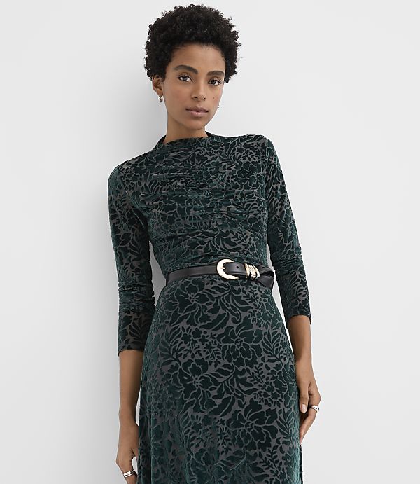 A woman wearing a dark green velvet floral dress with a black belt stands against a plain white background.