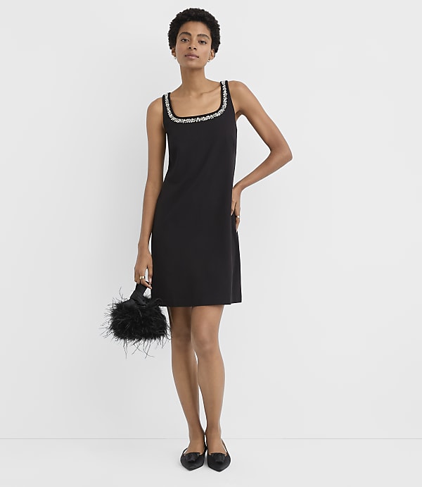 A woman wearing a black cocktail dress with embellished neckline and holding a feathery black bag stands against a plain white background.