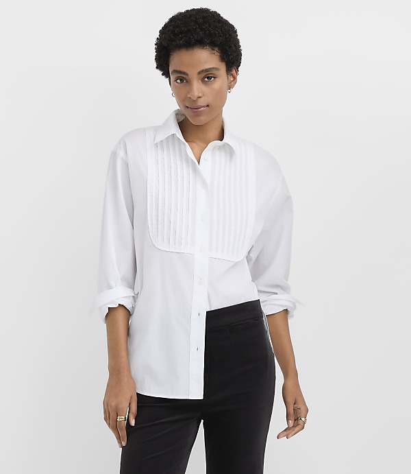 A woman wearing a white pleated button-down shirt and black trousers stands against a plain white background.