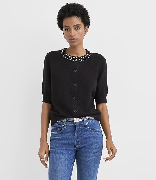 A woman wearing a black embellished cardigan and blue jeans stands against a plain light gray background.