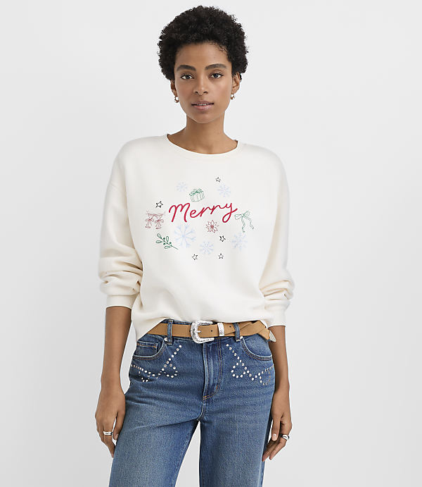 A woman wearing a white "Merry" sweatshirt and studded blue jeans stands against a plain white background.