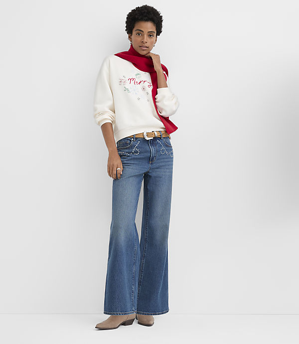 A woman wearing a white "Merry" sweatshirt, a red scarf, and wide-leg blue jeans stands against a plain white background.