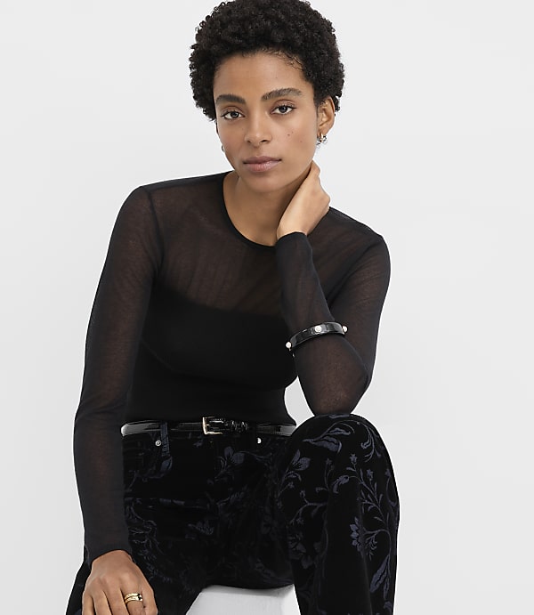 A woman wearing a sheer black long-sleeved top and floral velvet trousers poses against a plain white background.