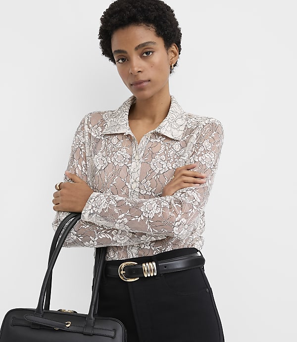 A woman wearing a floral lace blouse and black trousers stands with her arms crossed against a plain white background.
