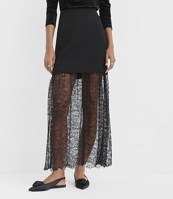 A person wears a black skirt with a sheer lace lower half and black pointed-toe flats against a plain white background.