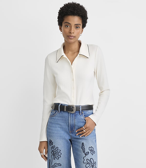 A woman in a cream-colored ribbed shirt and blue jeans with embroidered flowers stands against a plain white background.