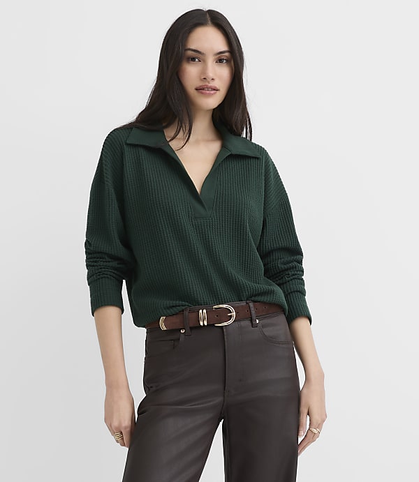A woman wearing a dark green ribbed collared top and brown leather pants stands against a plain white background.