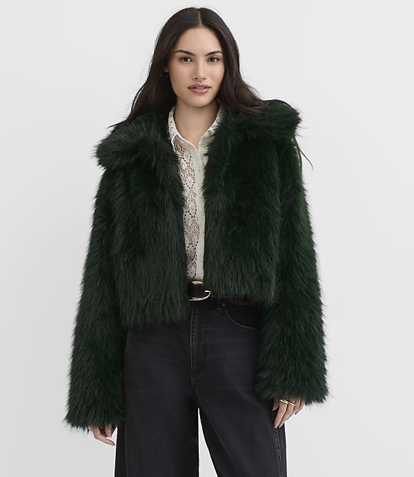 A woman wearing a dark green faux-fur jacket, a white lace blouse, and black jeans stands against a plain white background.
