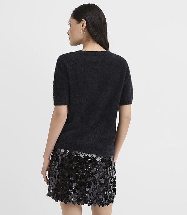 A woman wearing a fuzzy black short-sleeved top and a sequined black skirt stands with her back to the camera against a plain light gray background.