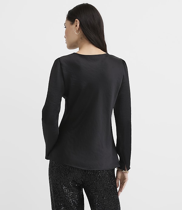 A woman wearing a black long-sleeved blouse and sequined black pants stands with her back to the camera against a plain white background.