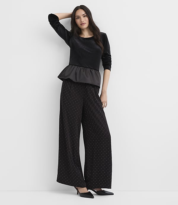 A woman wearing a black velvet peplum top and wide-leg polka-dot trousers stands against a plain white background.