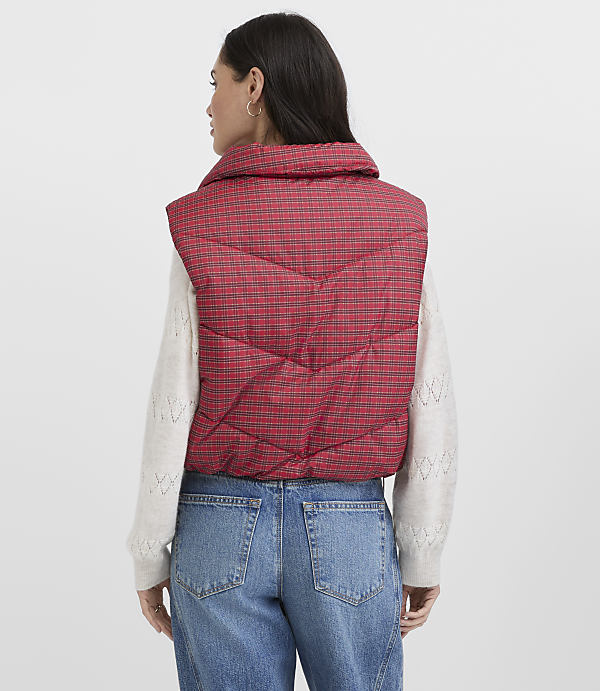 A woman wearing a red plaid puffer vest, a white sweater, and blue jeans stands against a plain light gray background.