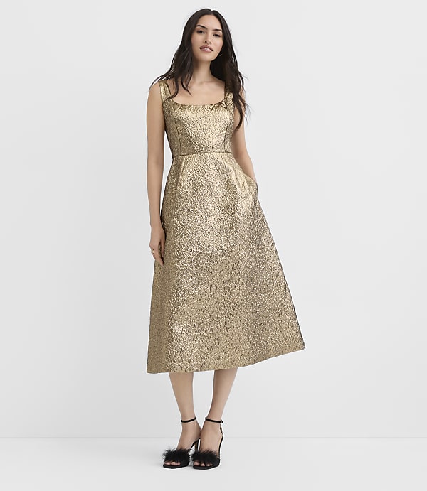 A woman wearing a shimmering gold midi dress and black feathered heels stands against a plain white background.