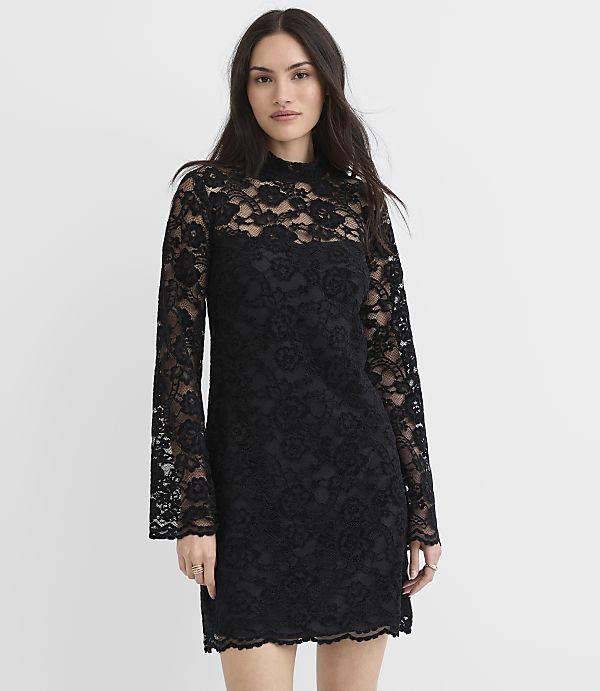 A woman wearing a black lace long-sleeved mini dress stands against a plain white background.