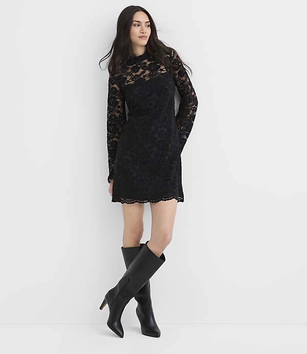 A woman wearing a black lace dress and black boots leans against a plain white wall.