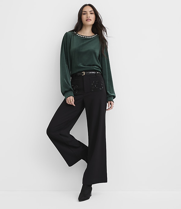 A woman wearing a green long-sleeved top and black wide-leg trousers stands against a plain white background.