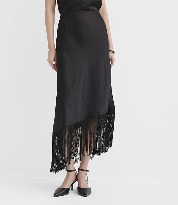 A person wears a black satin midi skirt with a fringed hem and black pointed-toe heels against a plain white background.