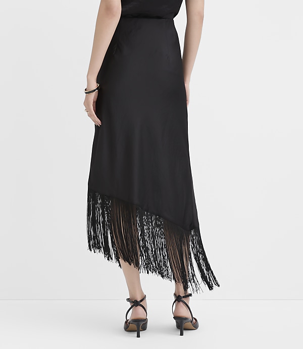 A person wearing a black midi dress with a fringed hem and black strappy heels stands against a plain white background.