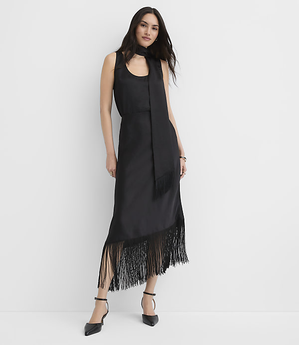 A woman wearing a black sleeveless midi dress with fringe detailing stands against a plain white background.