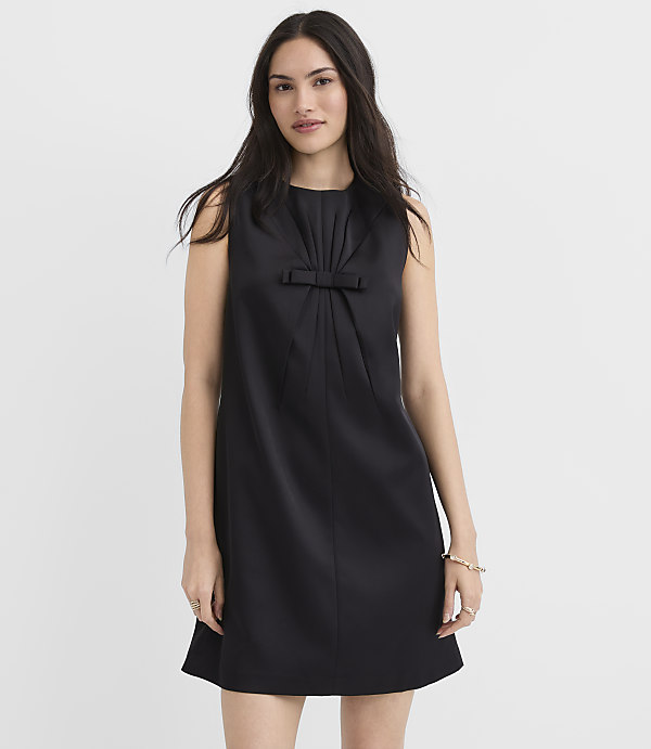 A woman wearing a sleeveless black dress with a bow detail stands against a plain white background.