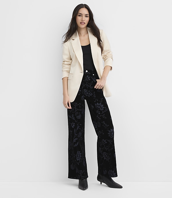 A woman wearing a cream blazer and floral black trousers stands against a plain white background.