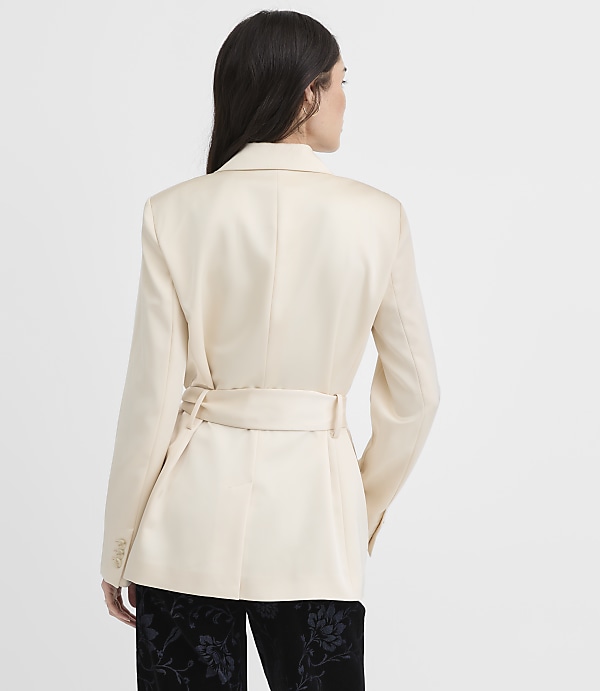 A woman wearing a cream-colored belted blazer and black floral trousers stands against a plain white background.