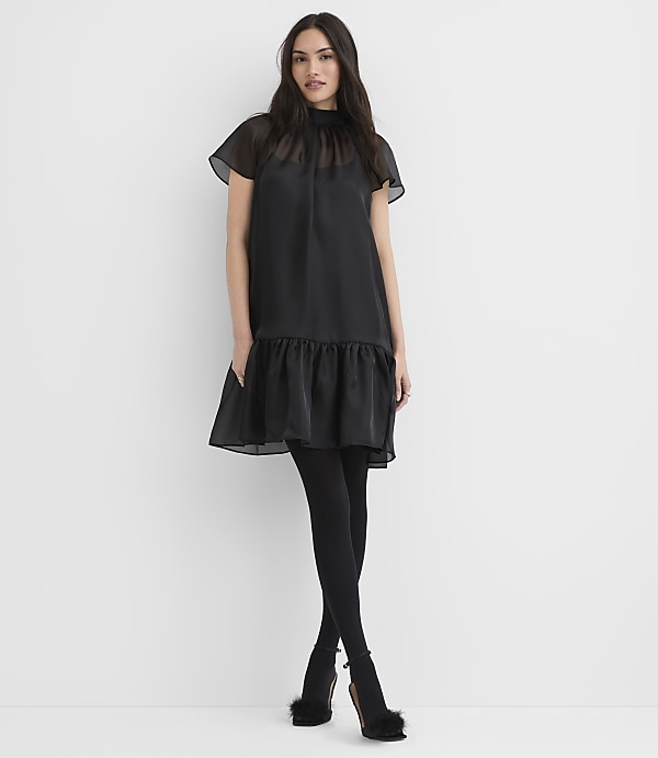 A woman wearing a black dress, tights, and fuzzy heels stands against a plain white background.