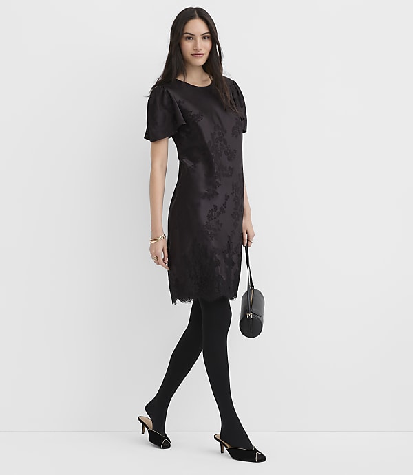 A woman wearing a black floral dress, tights, and heels stands against a plain white background.