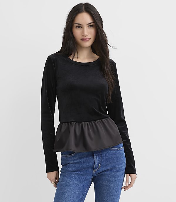 A woman wearing a black long-sleeved velvet top with a satin peplum waist and blue jeans stands against a plain white background.