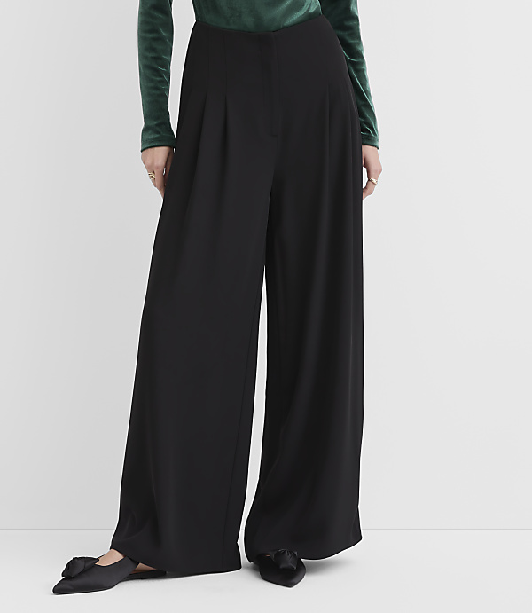 A person wearing a green velvet top and wide-leg black trousers stands against a plain white background.