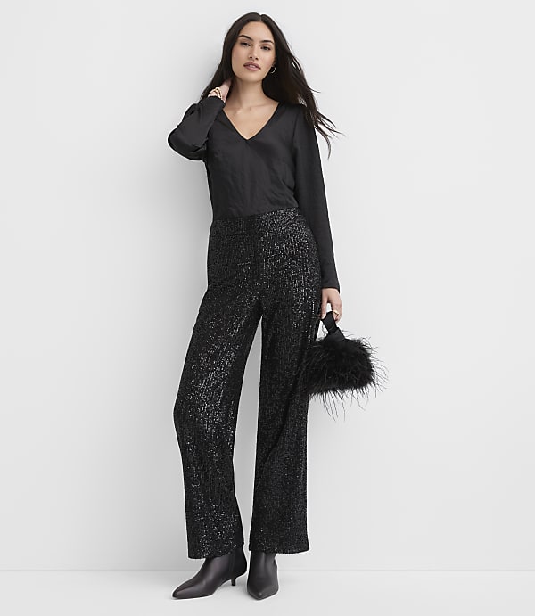 A woman wearing a black V-neck top and shimmering black wide-leg trousers stands against a plain white background.