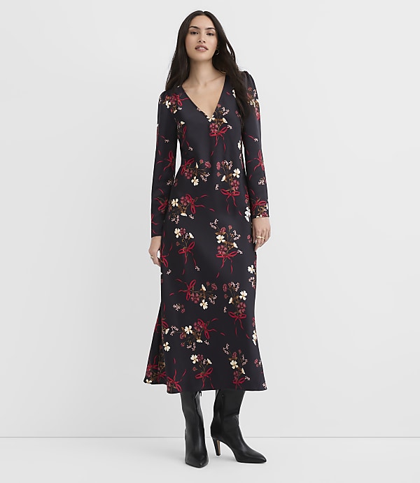 A woman wearing a long-sleeved black floral dress and black boots stands against a plain white background.